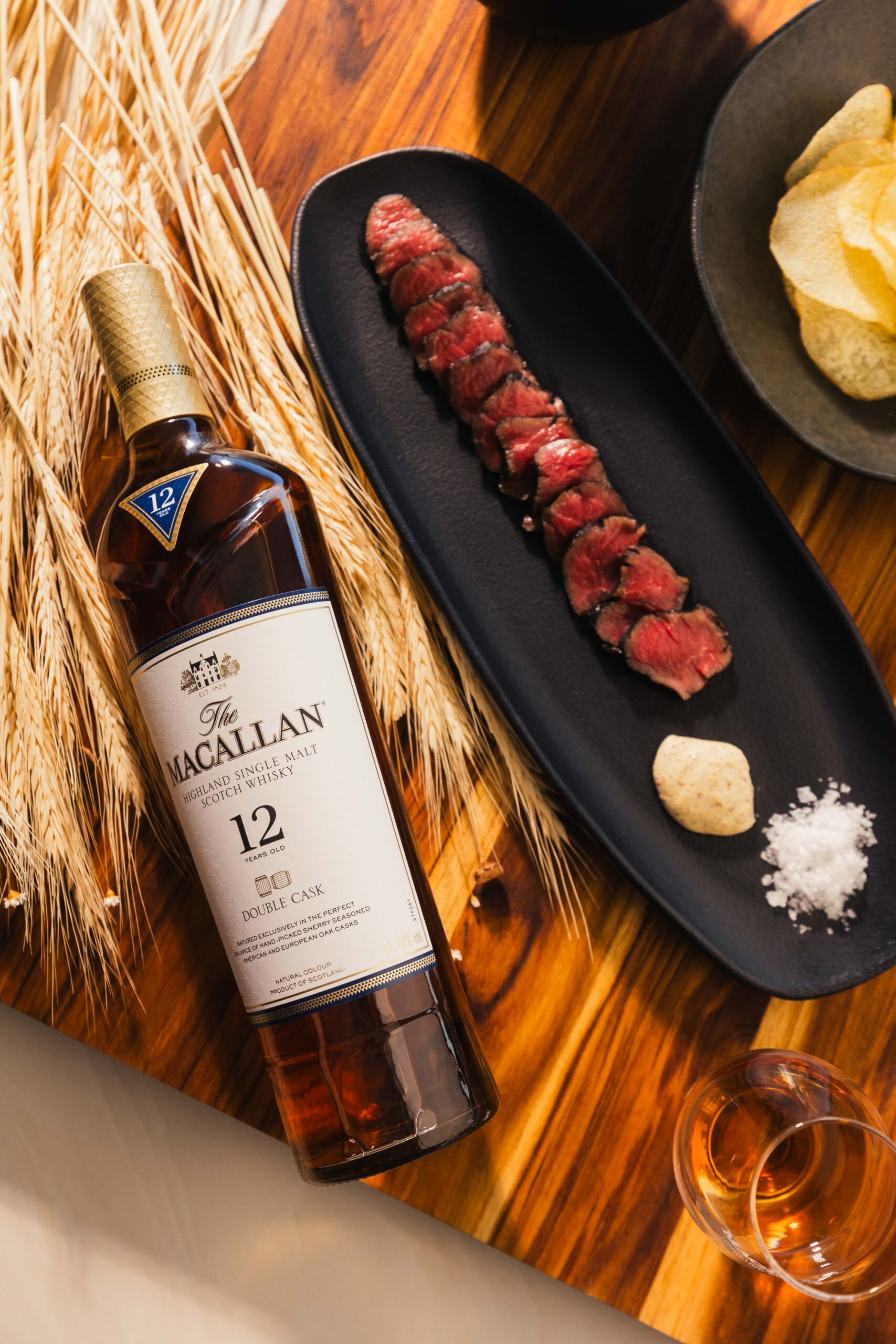 Vista superior de uma garrafa de uísque Macallan 12 anos e um prato de tartare de carne sobre mesa de madeira rusticada, foto por Lion Audiovisual.