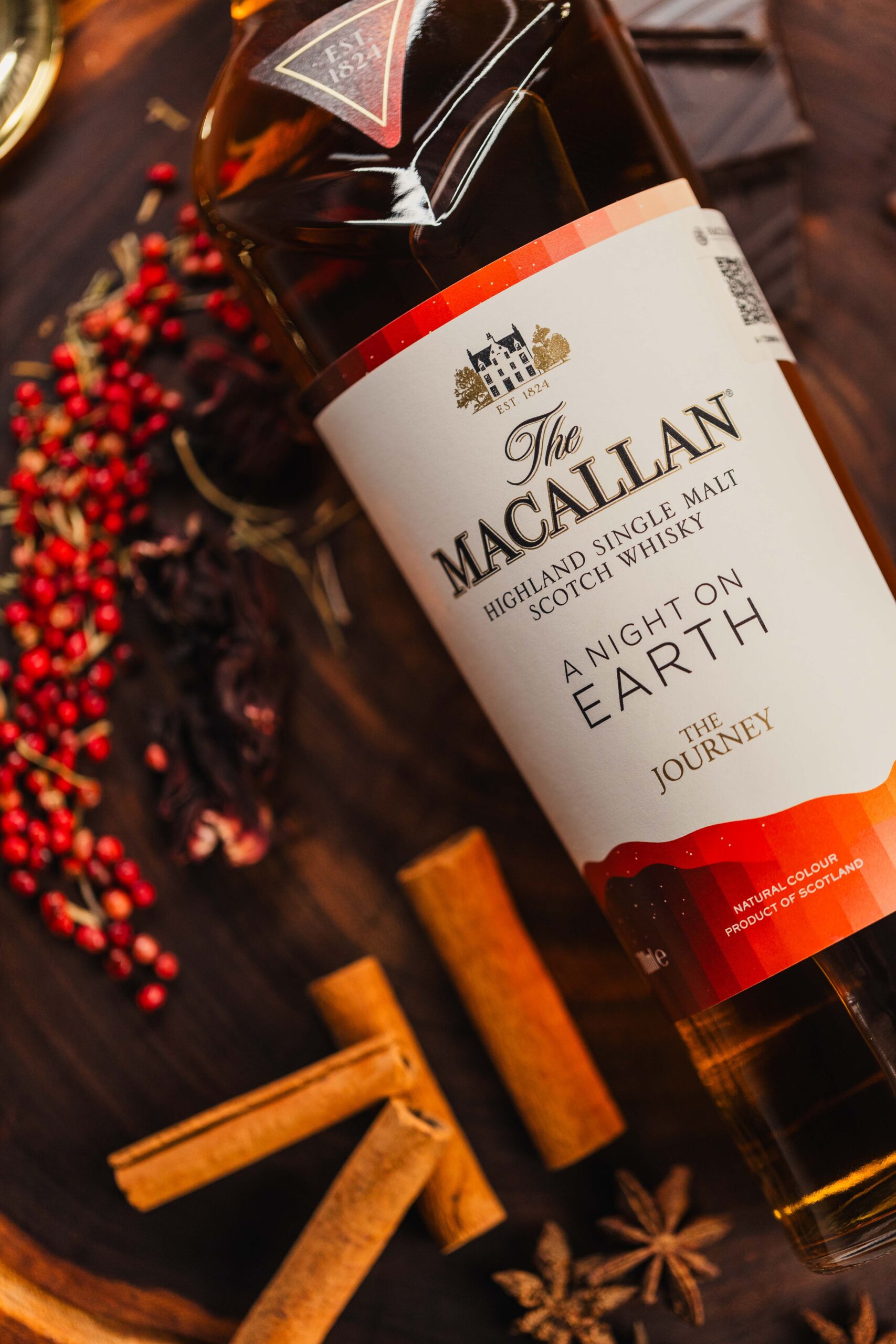 Garrafa de whisky The Macallan A Night on Earth sobre fundo de madeira com especiarias e frutas vermelhas.