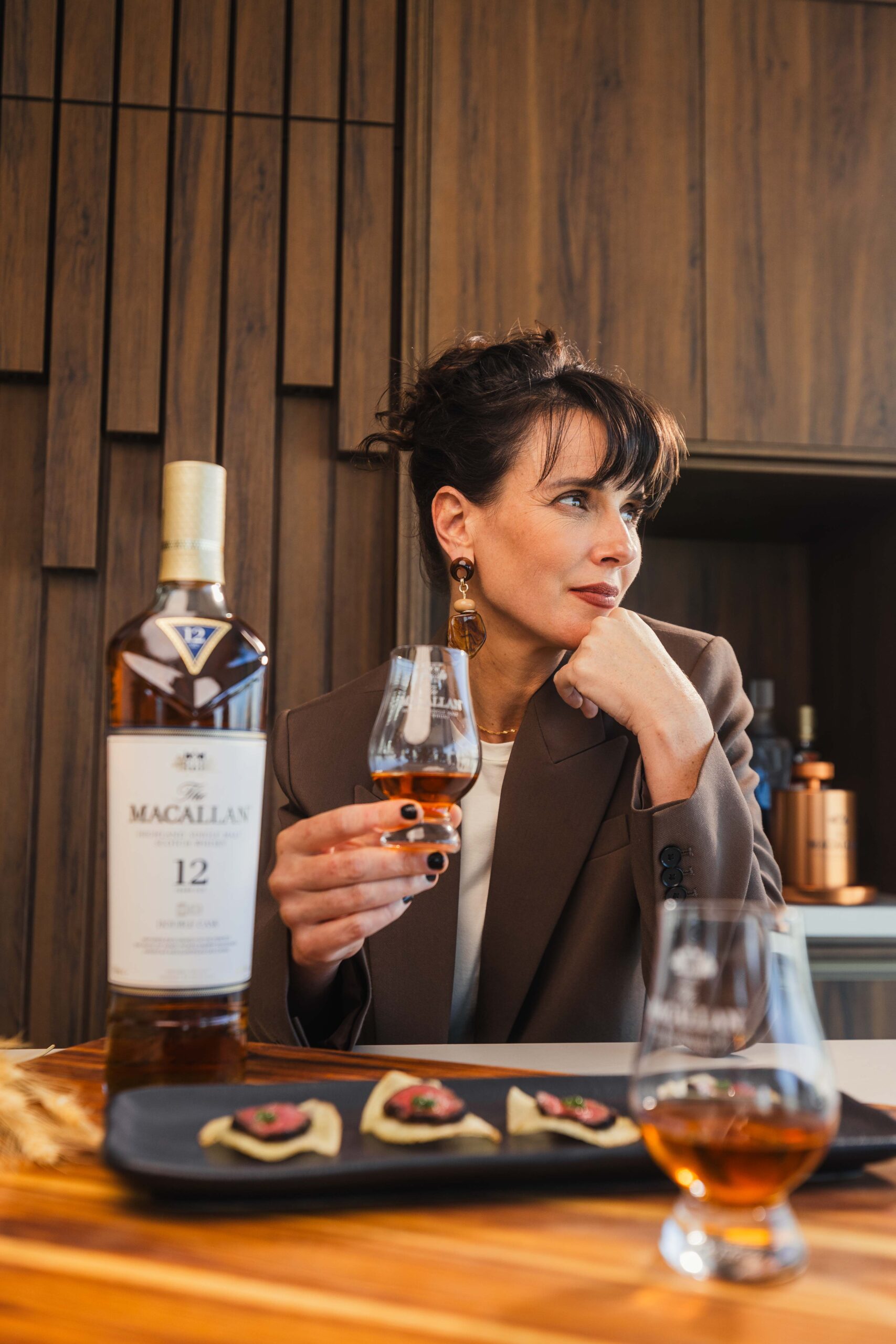 Chef Helena Rizzo observando taça de whisky com garrafa de The Macallan 12 Anos e prato harmonizado na mesa.