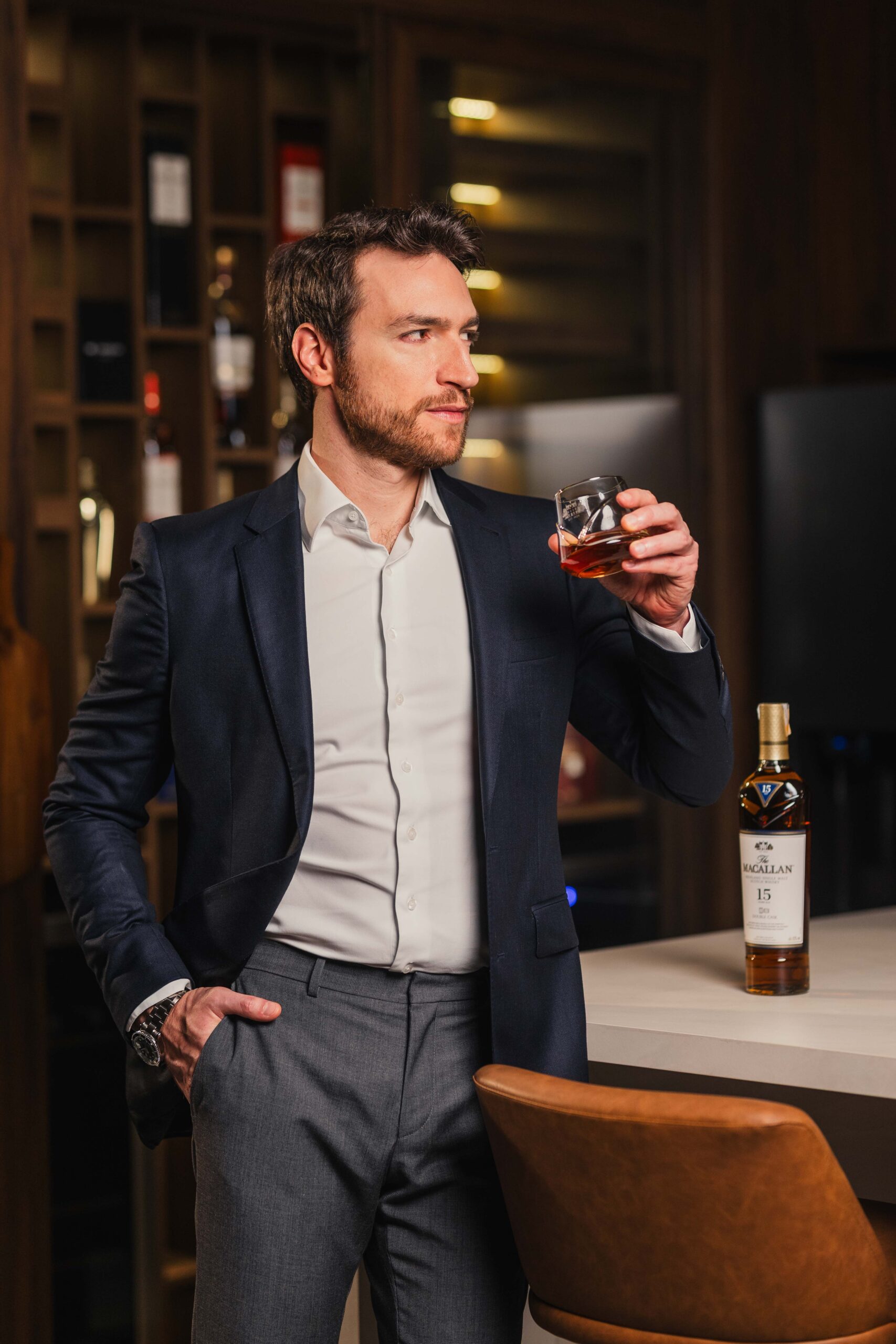 Gianpaolo Morselli, embaixador da The Macallan, vestindo terno e segurando copo de whisky em bar elegante.