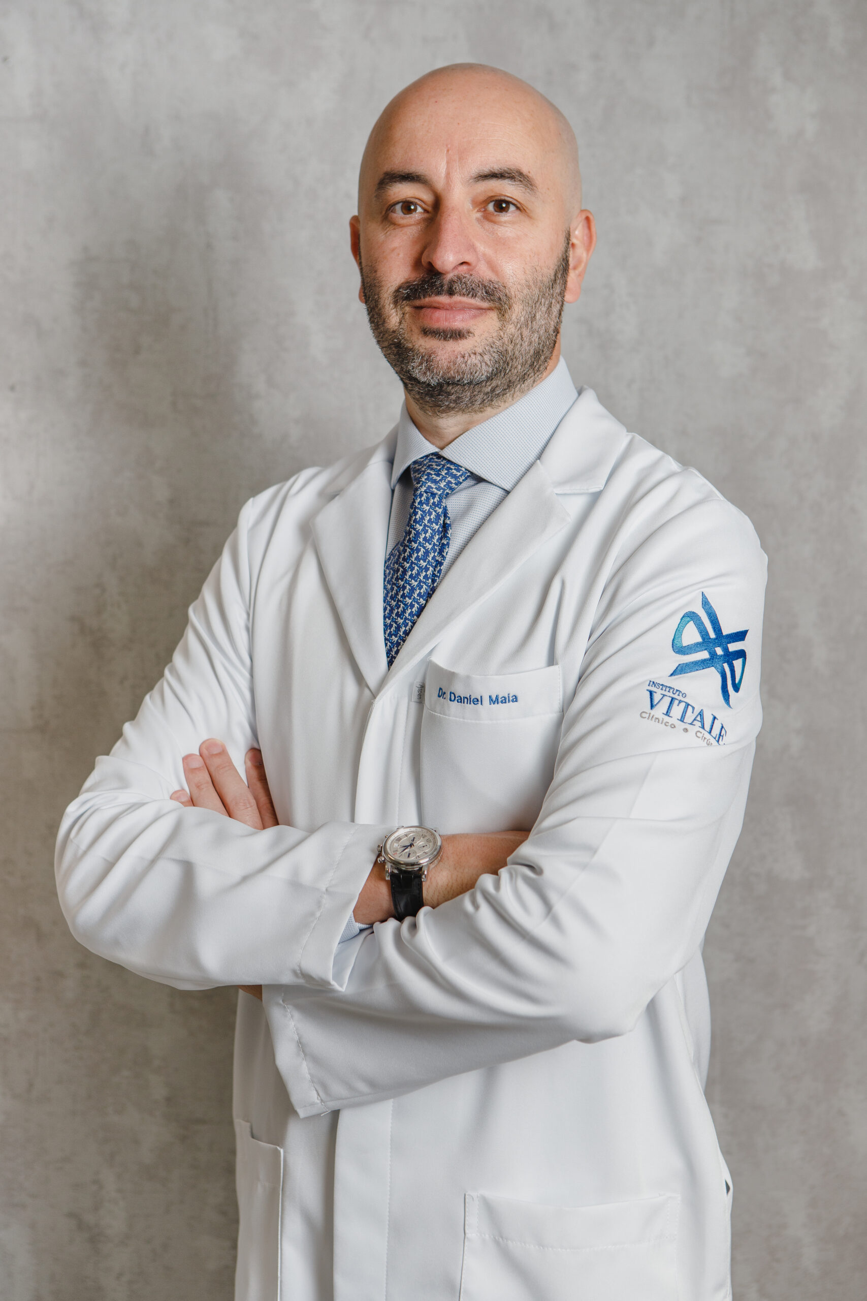 Médico do Instituto Vitale de jaleco branco com braços cruzados sorrindo em fundo cinza.