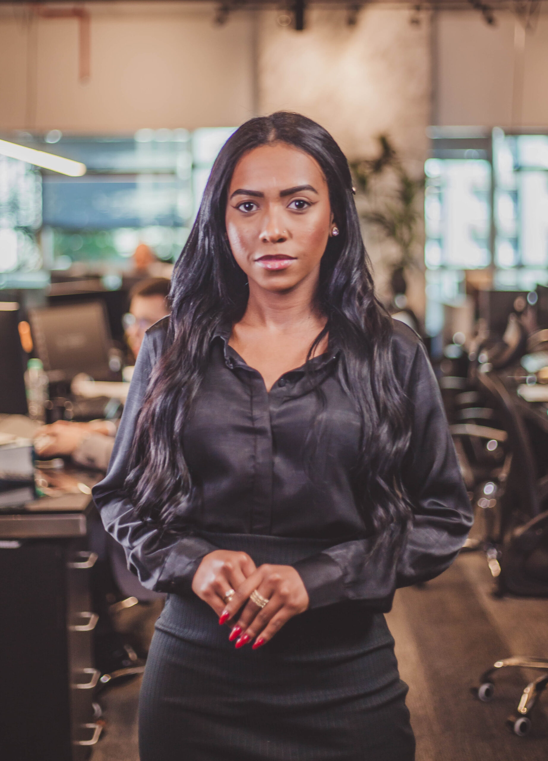 Mulher negra de cabelos longos e ondulados, usando blusa acetinada preta e saia, de pé em um escritório moderno.